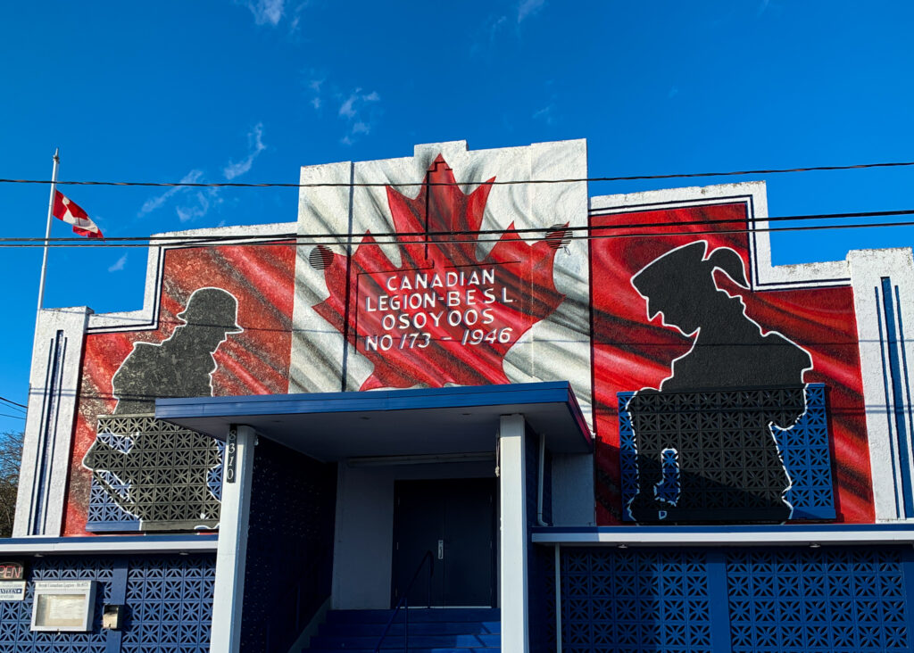 Photo of Royal Canadian Legion Hall in Osoyoos with a large Canadian flag painted on the front, flanked by two soldier silhouettes. 